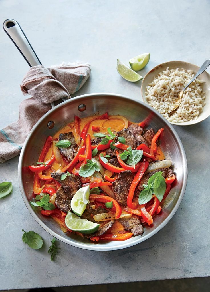 a pan filled with meat and vegetables next to rice, lime wedges and cilantro