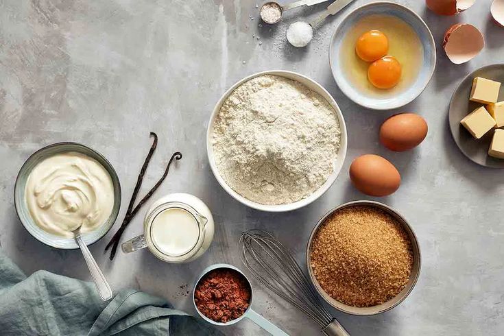 an overhead view of various ingredients and utensils on a gray background, including eggs, flour, butter, sugar, and other items