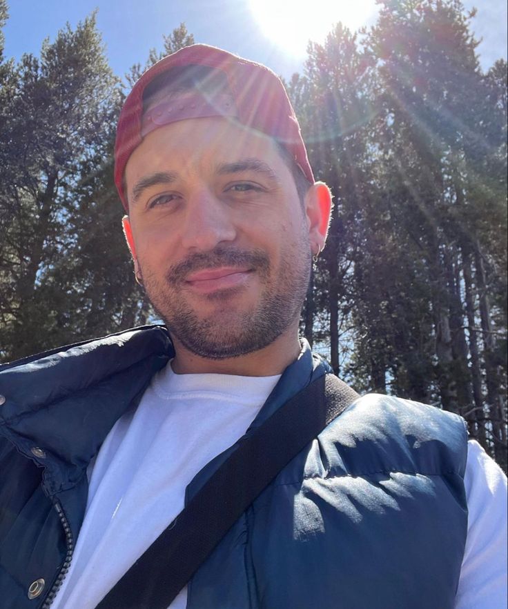 a man wearing a red hat standing in front of pine trees with the sun shining on him