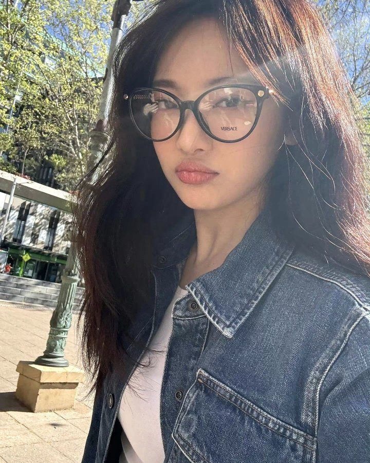 a woman wearing glasses standing in front of a street light with trees and buildings behind her