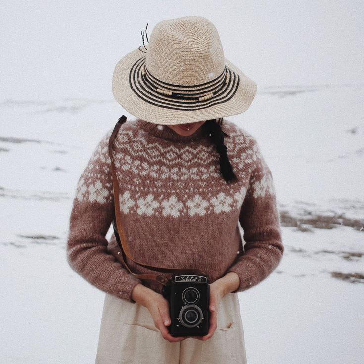 a woman wearing a hat and holding a camera in her hands while standing on snow covered ground