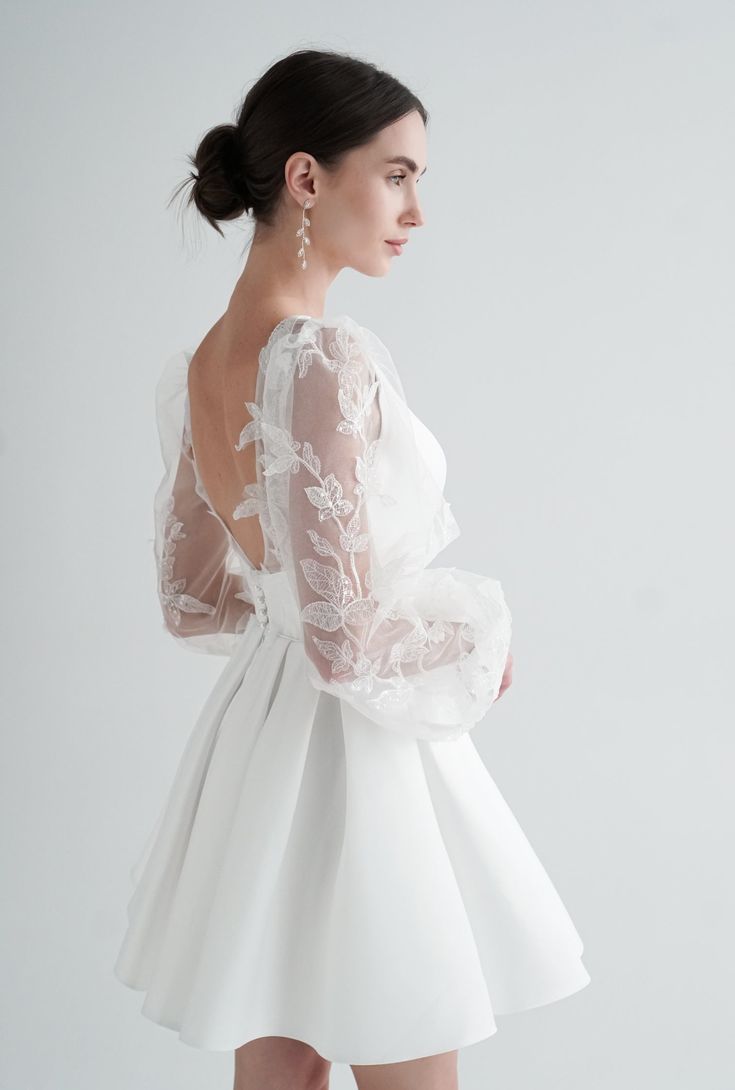 a woman wearing a white dress with long sleeves and lace on the back, standing in front of a white background