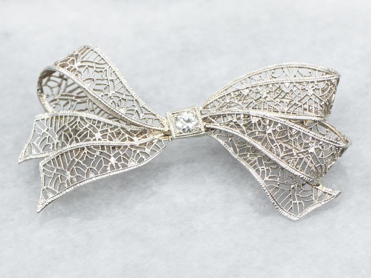 Add a touch of elegance and whimsy to any outfit with this bow brooch. The delicate filigree detailing and sparkling European Cut diamond make this brooch a timeless statement piece. Perfect for any occasion!Metal: 14K White GoldGem: European Cut Diamond .10 Carats, SI1 in Clarity, I in ColorGem Measurements: 3.2 mm, RoundMeasurements: 44 x 23 mmMarks: "X14K" Stamped on the pin guard Diamond Filigree Brooches As Gift, Elegant Wedding Jewelry With Decorative Bow, Silver Bow Brooch For Wedding, Elegant White Gold Brooch With Intricate Design, Elegant White Gold Brooches With Intricate Design, Wedding Brooch With Intricate Diamond Design, Wedding Diamond Brooch With Intricate Design, Vintage Wedding Brooch With Bow Detail, Vintage Wedding Brooch With Bow