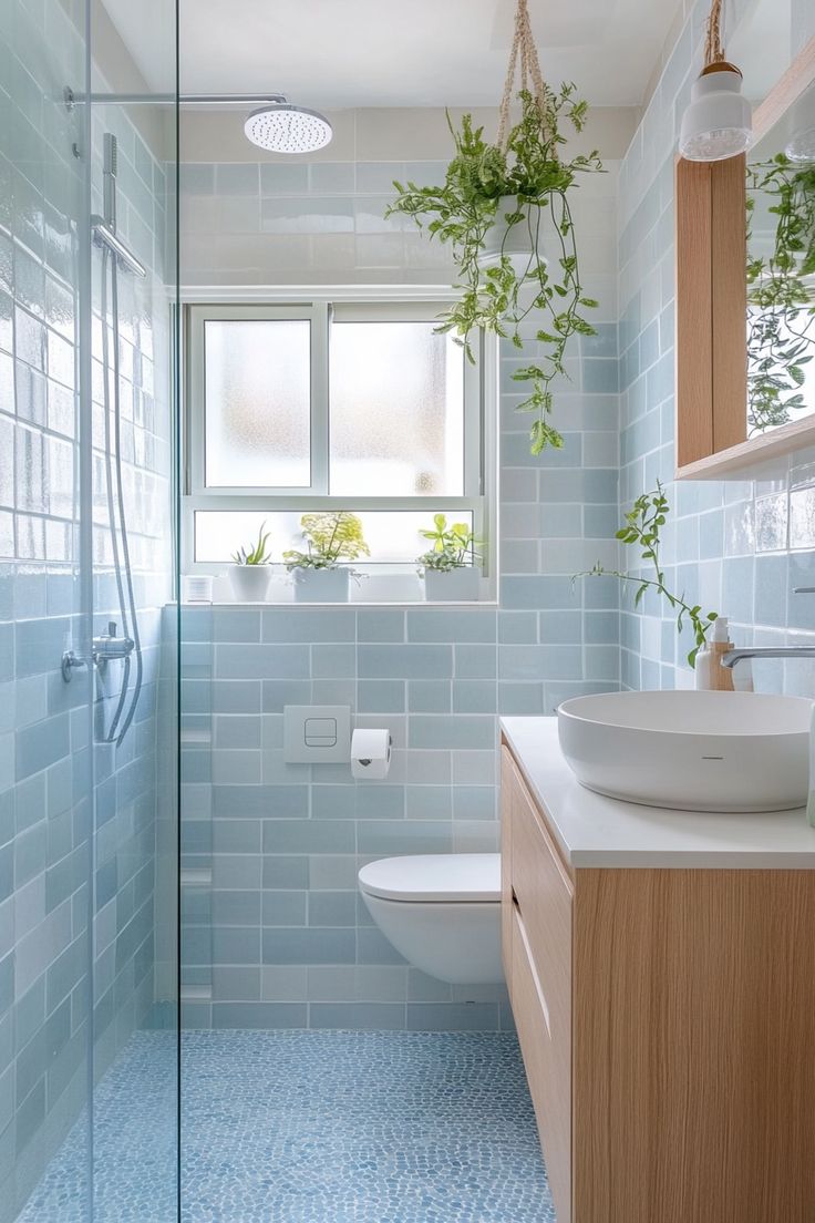 a bathroom with blue tile and wooden cabinetry is pictured in this image, there are plants hanging from the window above the sink