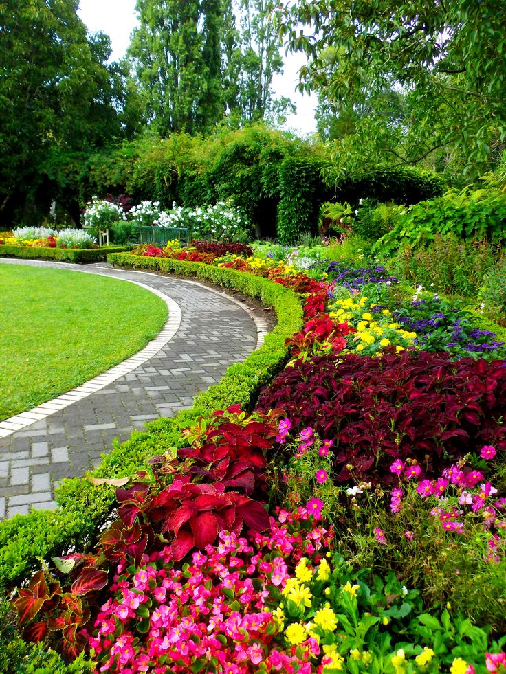 a garden with colorful flowers and greenery on the side of it, in front of a stone path