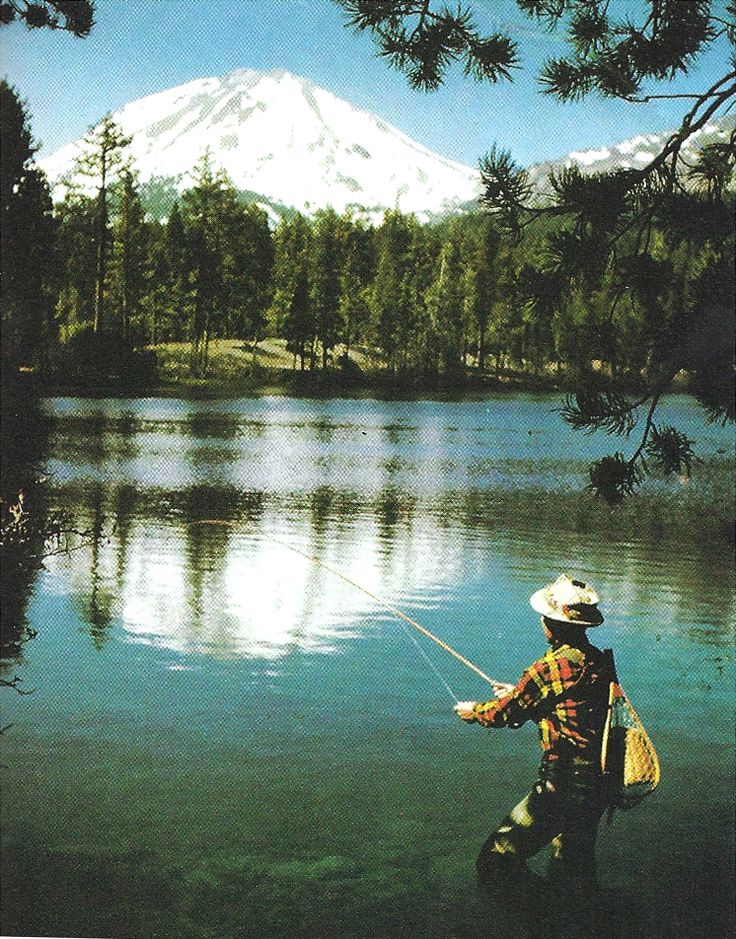 a man standing in the water holding a fishing pole and wearing a hat with a mountain in the background