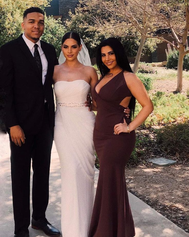 two women and a man standing next to each other on a sidewalk with trees in the background