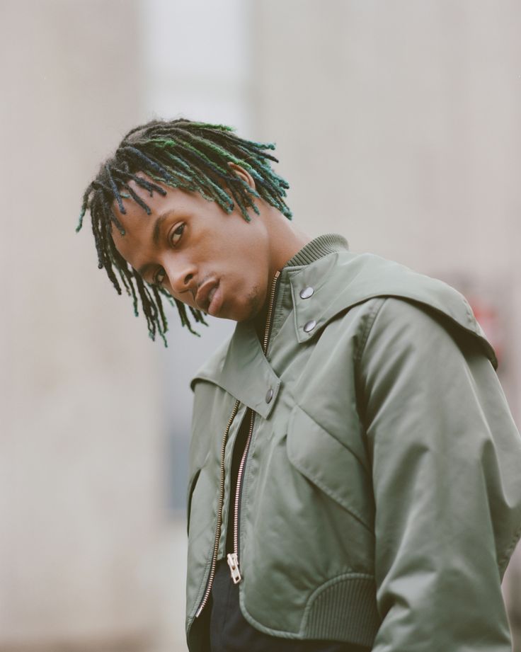 a young man with dreadlocks standing in front of a building wearing a green jacket