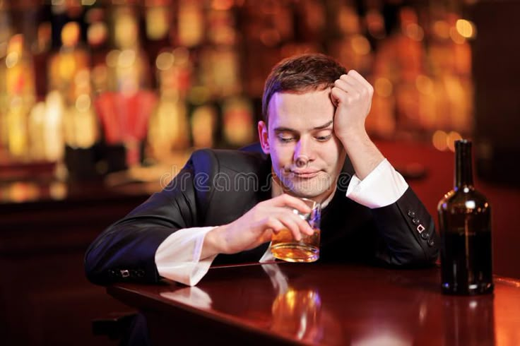 a man sitting at a table with a glass in front of him and bottles behind him