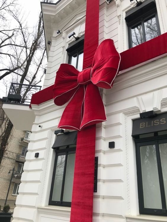 a large red bow tied to the side of a white building with windows and trees in the background