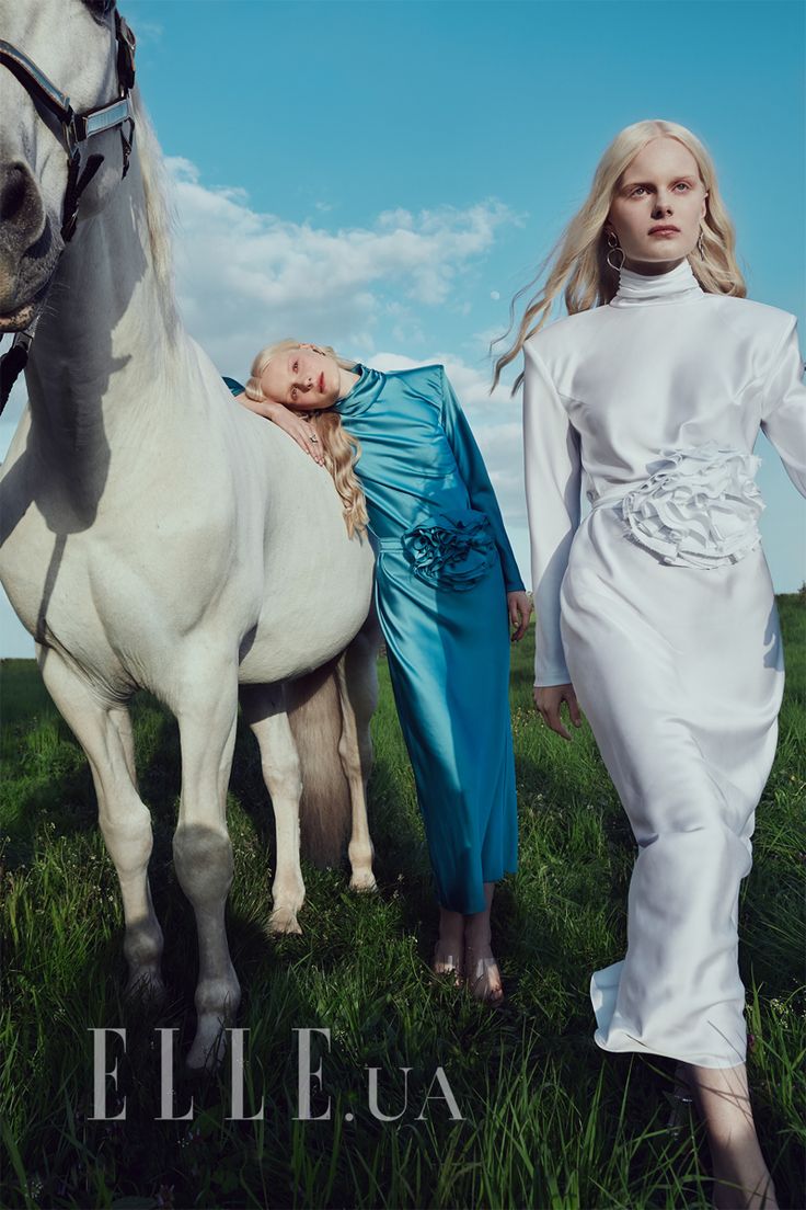 two women standing next to a white horse in a grassy field with the sky behind them