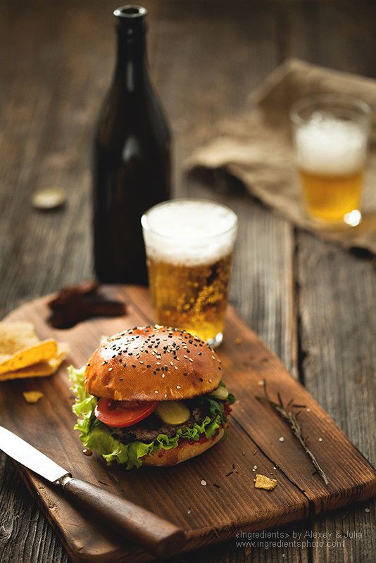 a hamburger and some chips on a wooden board next to two beer bottles with glasses