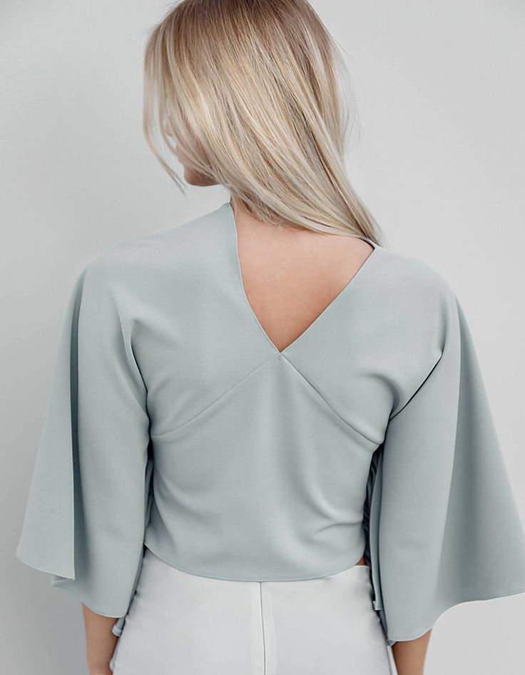 This blouse is constructed with 3/4 bell kimono sleeves, a high or lower asymmetrical V-neckline ending with a raglan seam. Features like a gathered front falling free garment on the sides made in cool gray shades with a blue undertone. The model wears size EU S Small: Bust: 89 cm, Waist: 74 cm 100% made in light blue scuba Modern V-neck Blouse For Spring, Modern Blue Long Sleeve Blouse, Modern Long Sleeve Blue Blouse, Elegant Blue Half Sleeve Top, Fitted Modern Top With 3/4 Sleeves, Fitted Tops With Draped Sleeves For Spring, Chic Blouse With Kimono Sleeves For Work, Chic Workwear Blouse With Kimono Sleeves, Modern Blue Blouse For Spring