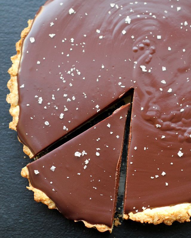 a chocolate tart with one slice missing from it on a black surface, next to a fork and knife