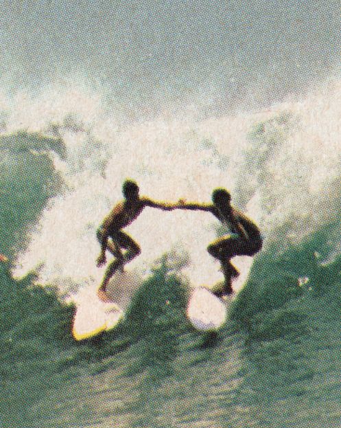 two people on surfboards in the middle of a wave, one holding another's hand