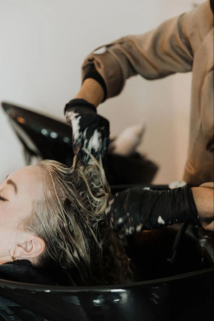 a woman getting her hair washed in a black bowl by a man with gloves on