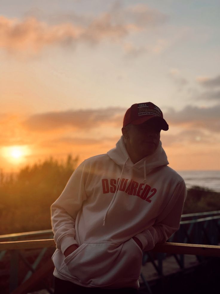 a man wearing a white hoodie and black pants standing in front of the sun