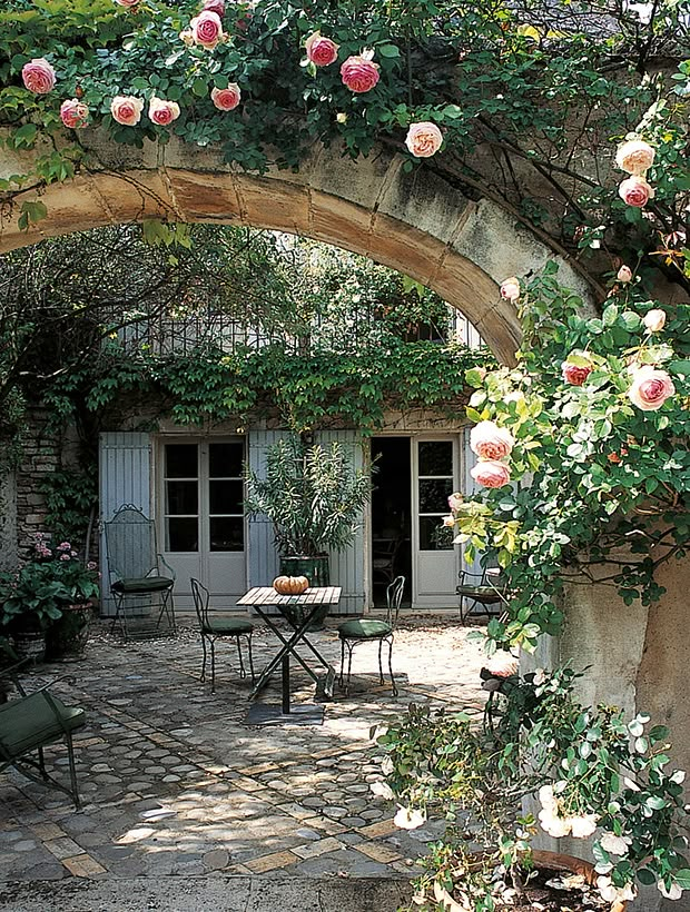 an archway with roses growing over it and tables in the foreground, surrounded by greenery