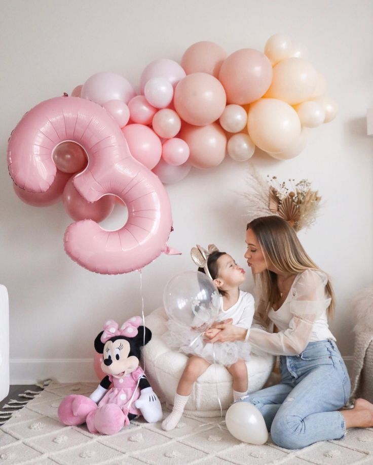 a woman holding a baby in front of balloons and a mickey mouse on the floor