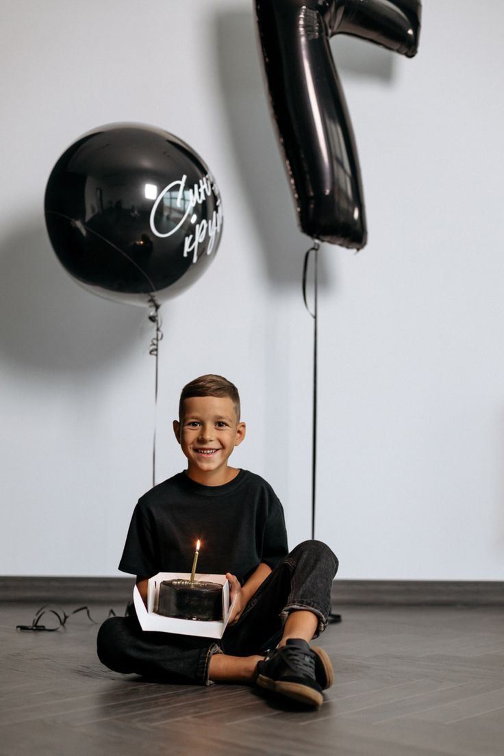 a young boy sitting on the floor with a cake in front of him and balloons behind him