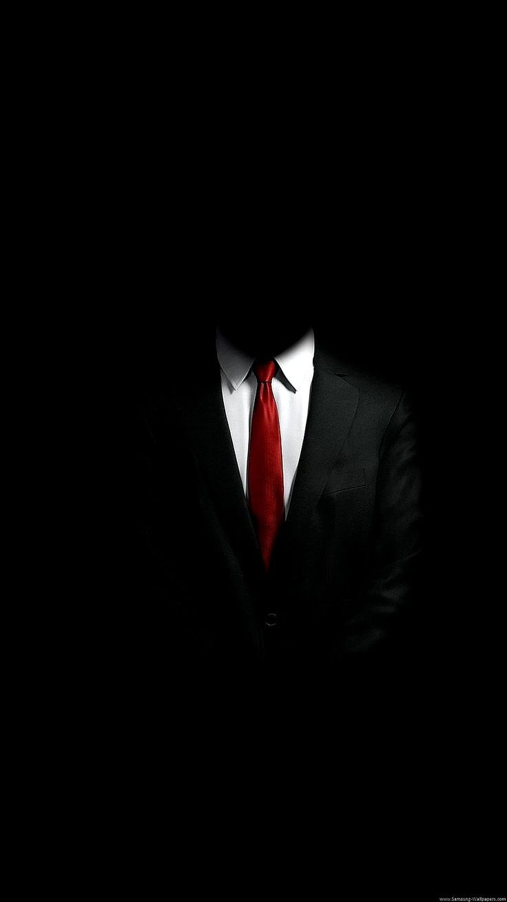 a man in a suit and red tie standing in the dark with his arms crossed