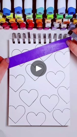 two hands holding a purple ribbon over a paper with hearts on it and markers in the background