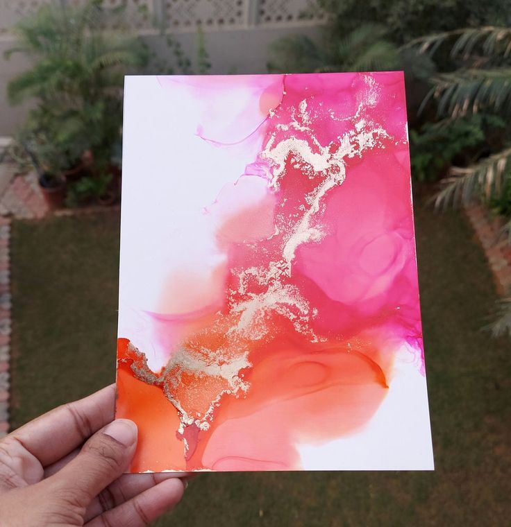 a hand holding up a piece of paper with pink and gold paint on it in front of a house