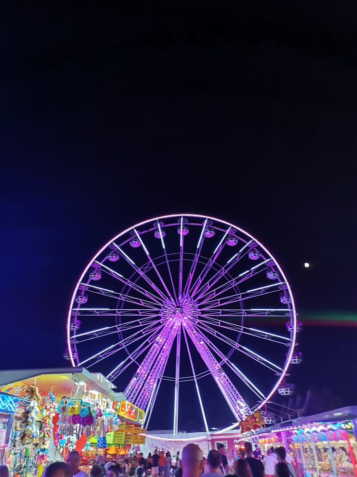 the ferris wheel is lit up at night with people walking around it and onlookers in the background