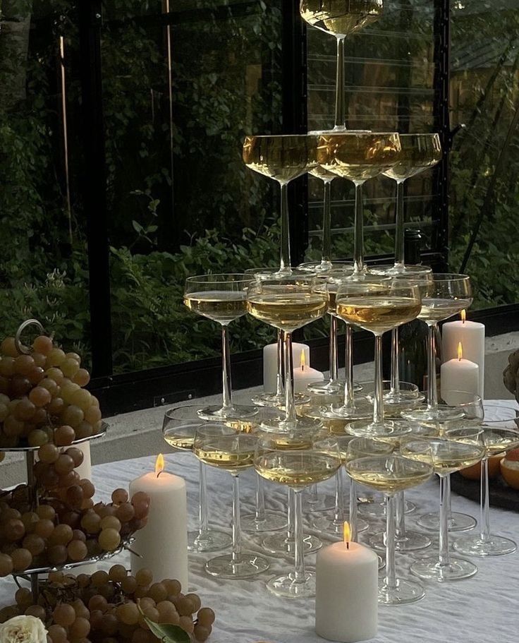 a table topped with lots of wine glasses filled with white wine next to candles and grapes