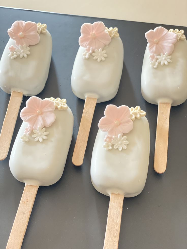 four pops with white frosting and pink flowers on them sitting on a black surface