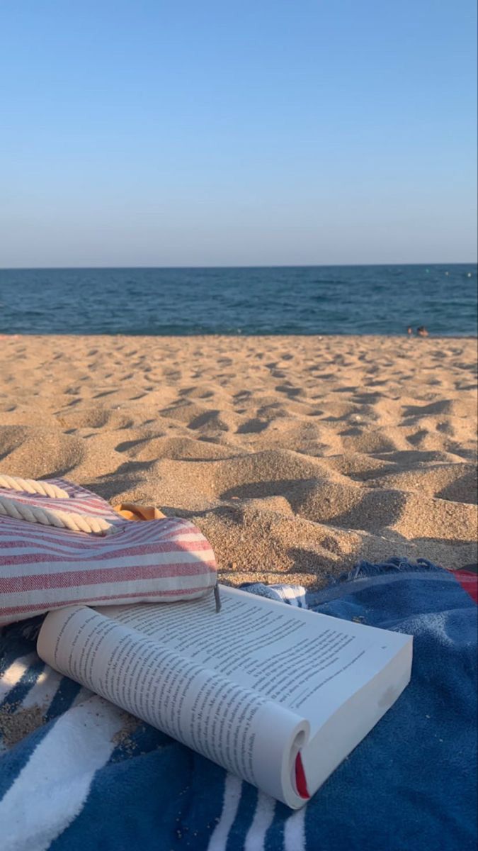 an open book sitting on top of a blue blanket next to the ocean and beach