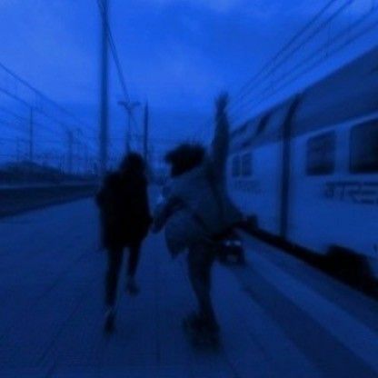 two people walking down a sidewalk next to a train on the tracks at night time