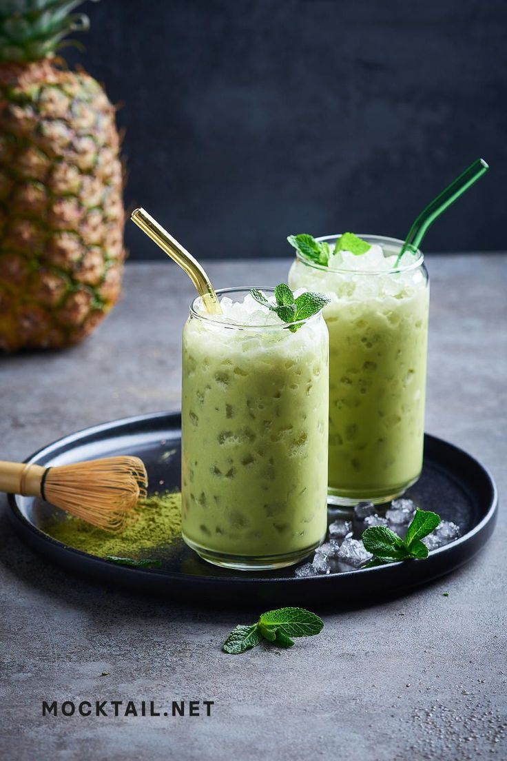 two glasses filled with green smoothie sitting on top of a plate next to a pineapple