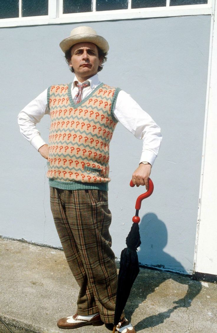 a man with an umbrella standing in front of a garage door wearing a vest and hat