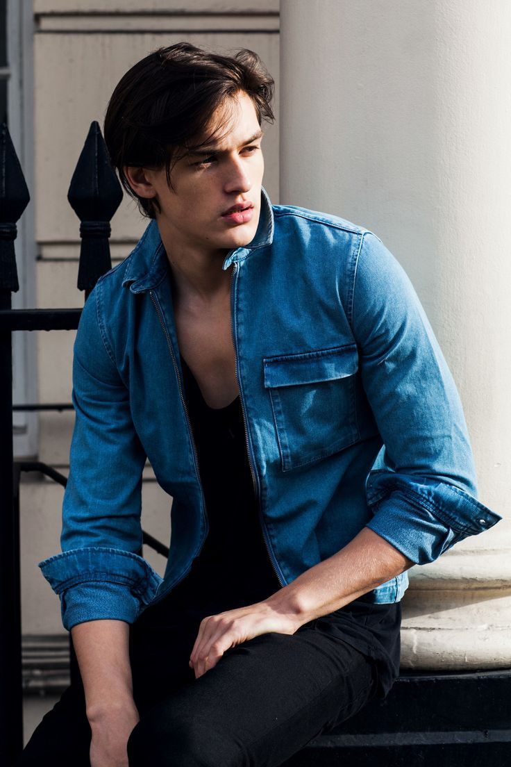 a young man sitting on the steps in front of a building wearing a denim shirt and black pants