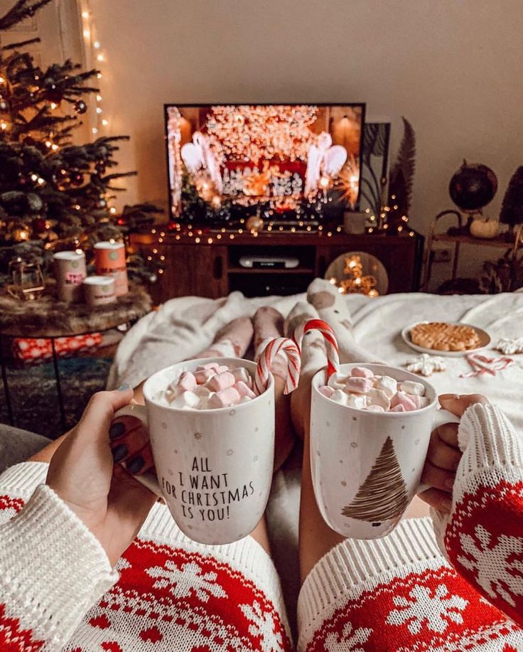 two people holding mugs with hot chocolate in front of a christmas tree and tv