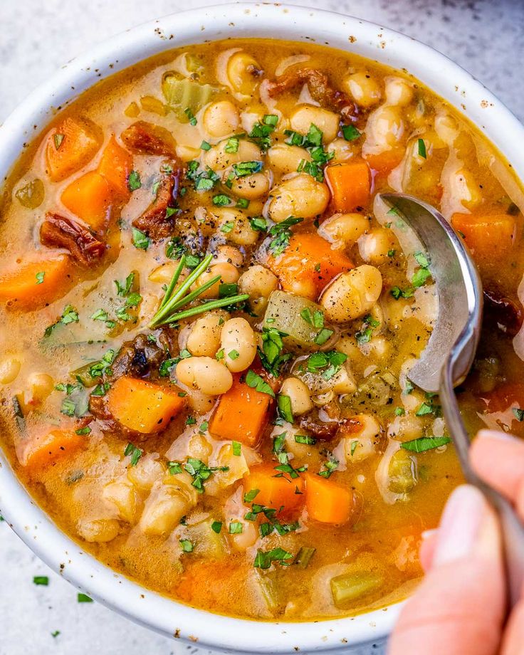 a person holding a spoon in a white bowl filled with beans, carrots and parsley