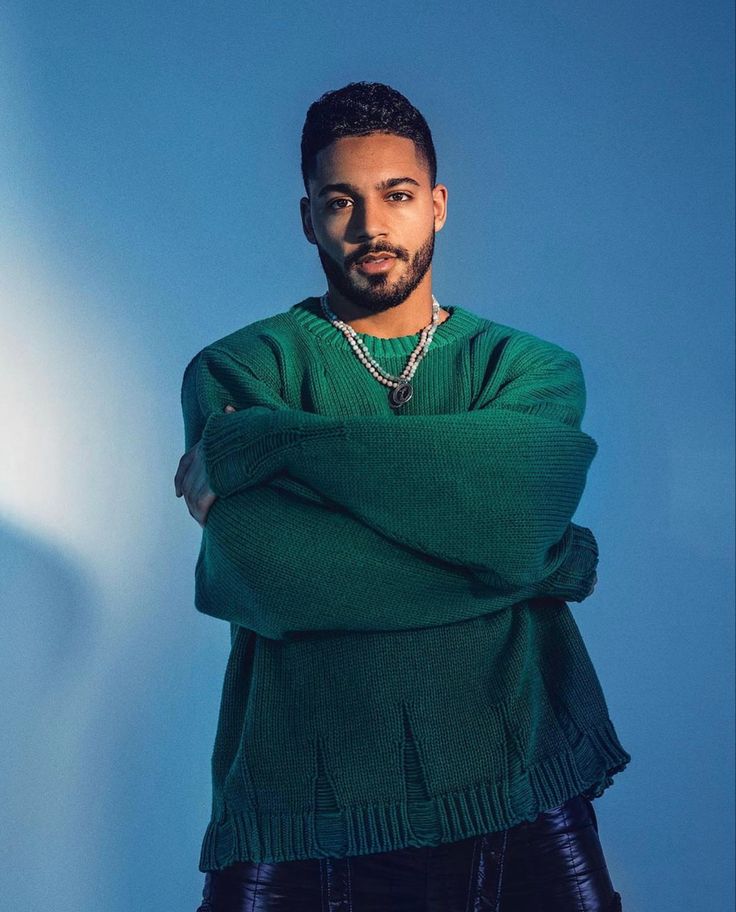 a man with his arms crossed standing in front of a blue wall wearing a green sweater