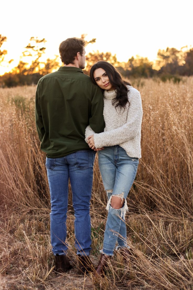 a young man and woman standing in tall grass holding each other's hands as the sun sets behind them