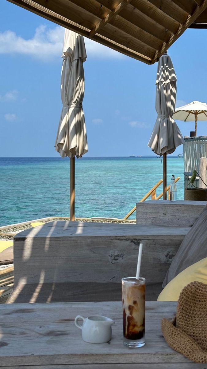 a cup of coffee sitting on top of a wooden table next to the ocean with umbrellas