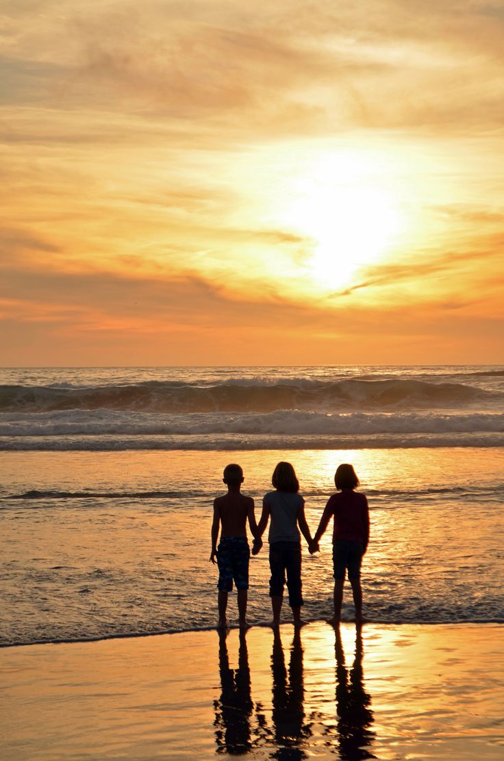 three people holding hands while standing on the beach at sunset or sunrise time with waves coming in