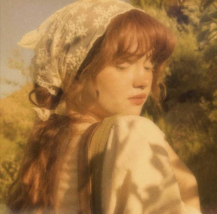 a woman with red hair wearing a white bonnet and looking off into the distance while standing in front of trees