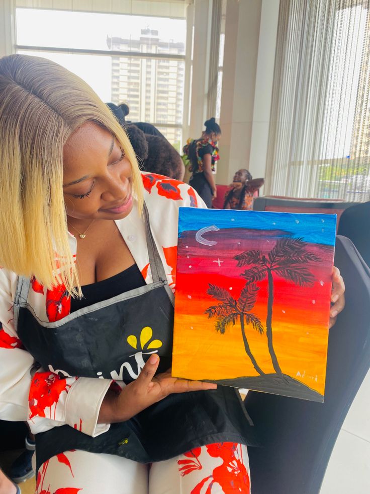 a woman holding a painting in her hands and looking down at it with palm trees painted on it