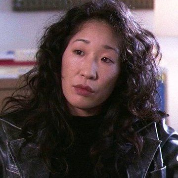 a woman with curly hair wearing a black leather jacket and looking at the camera while sitting in a chair