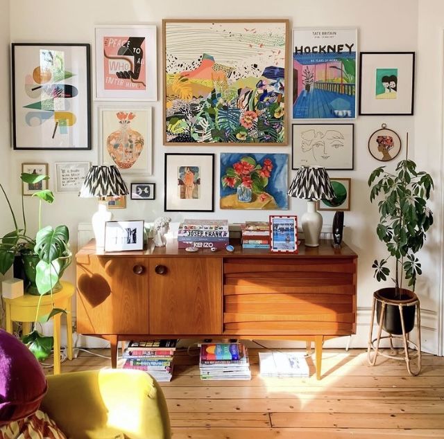 a living room filled with lots of art on the wall above a wooden dresser and colorful chairs