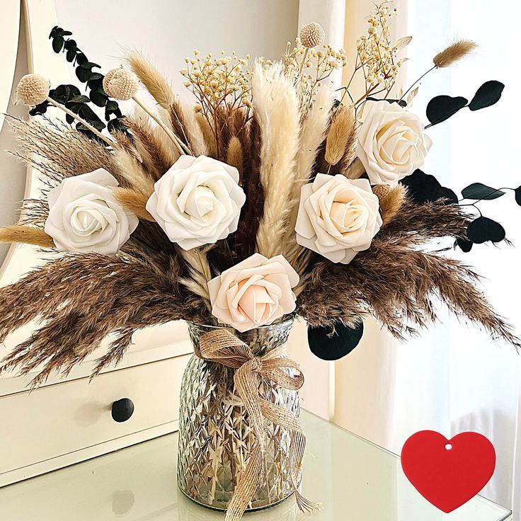 a vase filled with white roses and dry grass on top of a table next to a mirror