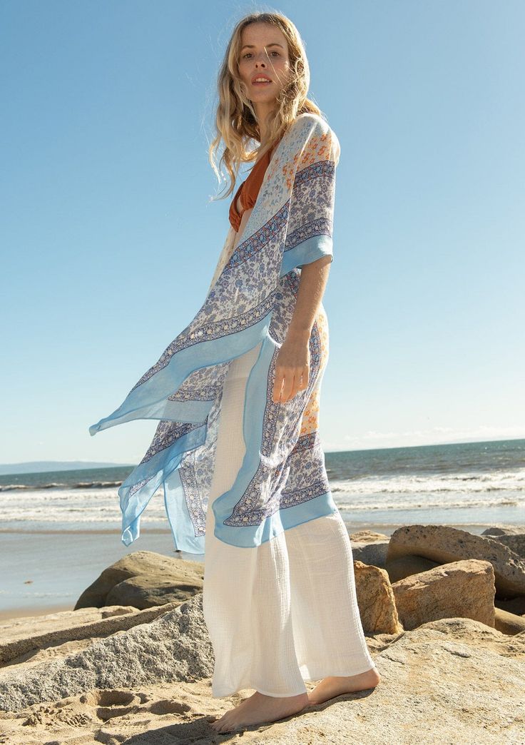 [Color: Ivory/Light Blue] A full body side facing image of a blonde model on the beach wearing a lightweight bohemian mid length kimono in an ivory and light blue mixed floral border print. With half length kimono sleeves Blue Boho Print Kimono For Spring, Blue Flowy Cover-up With Kimono Sleeves, Long Flowy Blue Kimono, Blue Flowy Long Kimono, Blue Boho Print Kimono For Beach Cover-up, Blue Boho Print Kimono For Beach, Bohemian Wrap Kimono For Daywear, Bohemian Summer Kimono With 3/4 Sleeves, White Boho Print Kimono For Spring