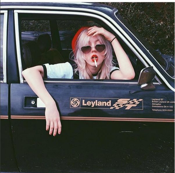 a woman with red lipstick sitting in the drivers seat of a black car wearing sunglasses