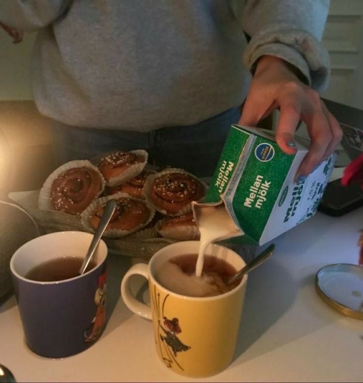 a person pours milk from a carton into two mugs filled with hot chocolate