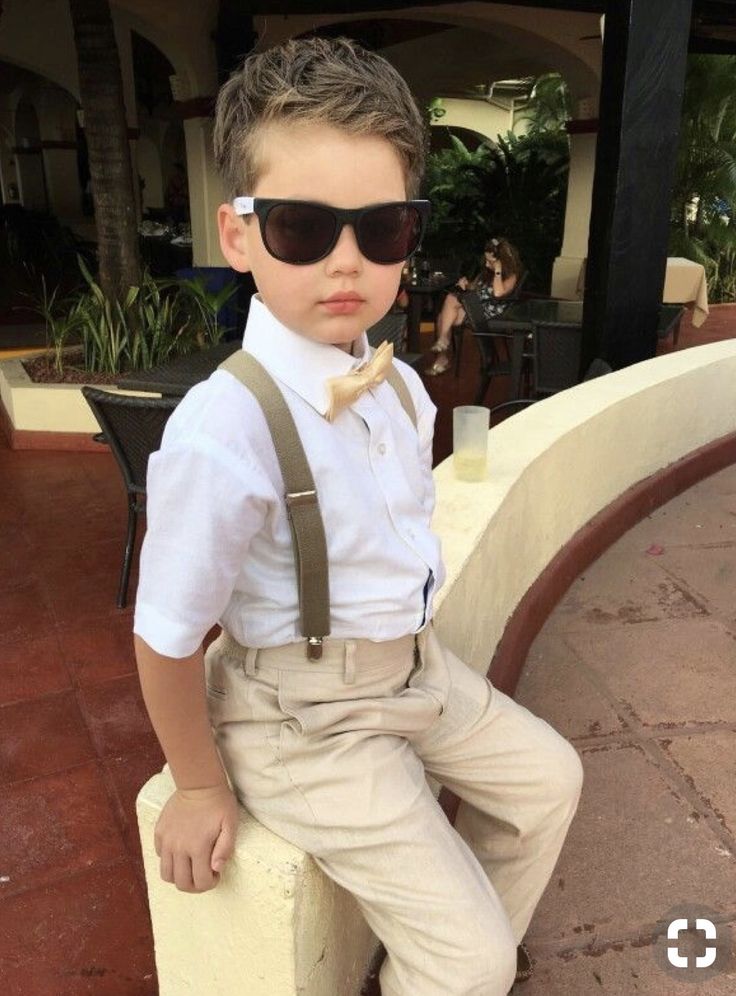 a young boy wearing sunglasses and suspenders sitting on a ledge